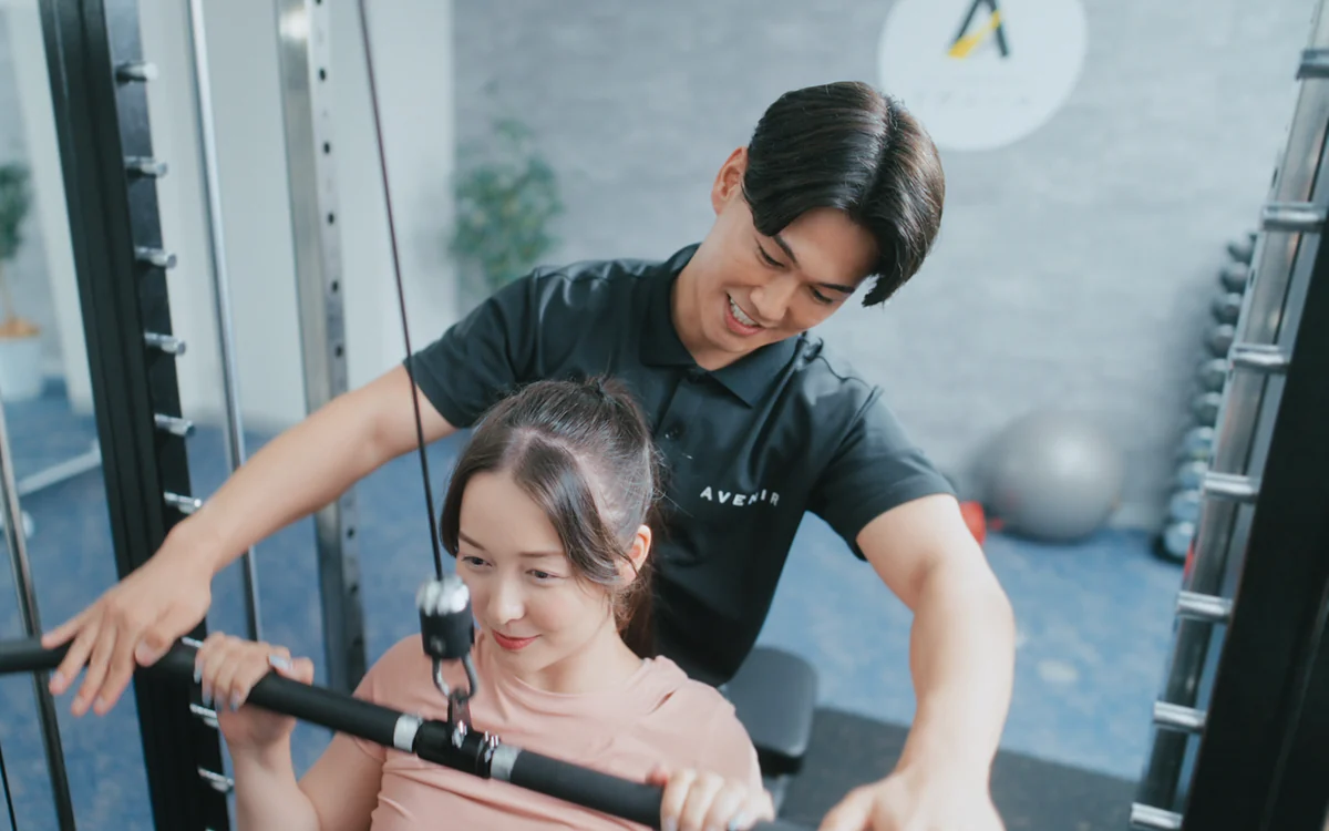 マシントレーニングをする女性と、そばで支えるトレーナー